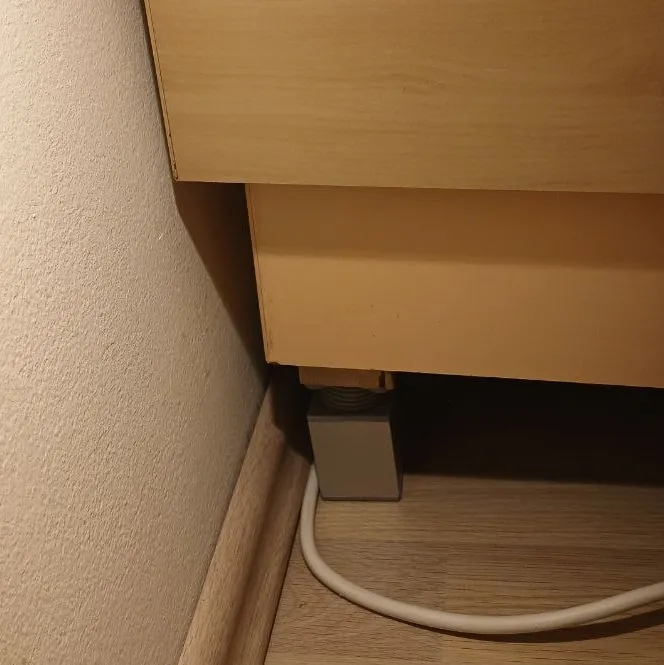 A grey plastic bed riser block placed under the wooden leg at the head of a bed, raising it several centimetres off the floor.